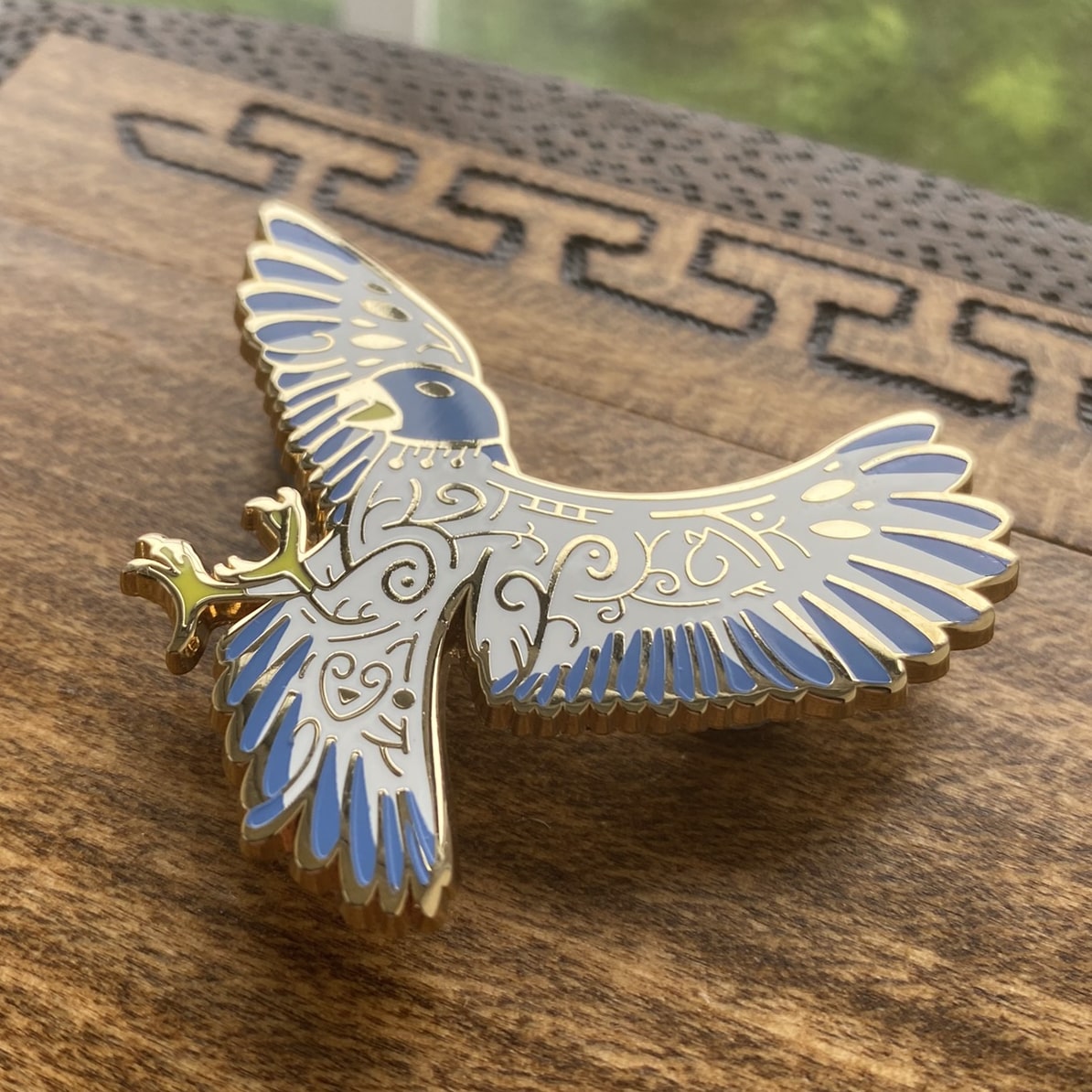 Gold-plated enamel pin of the Norse god Freyja. Pin contains knotwork and is depicted as a blue falcon. Angle shows off gold-plating.