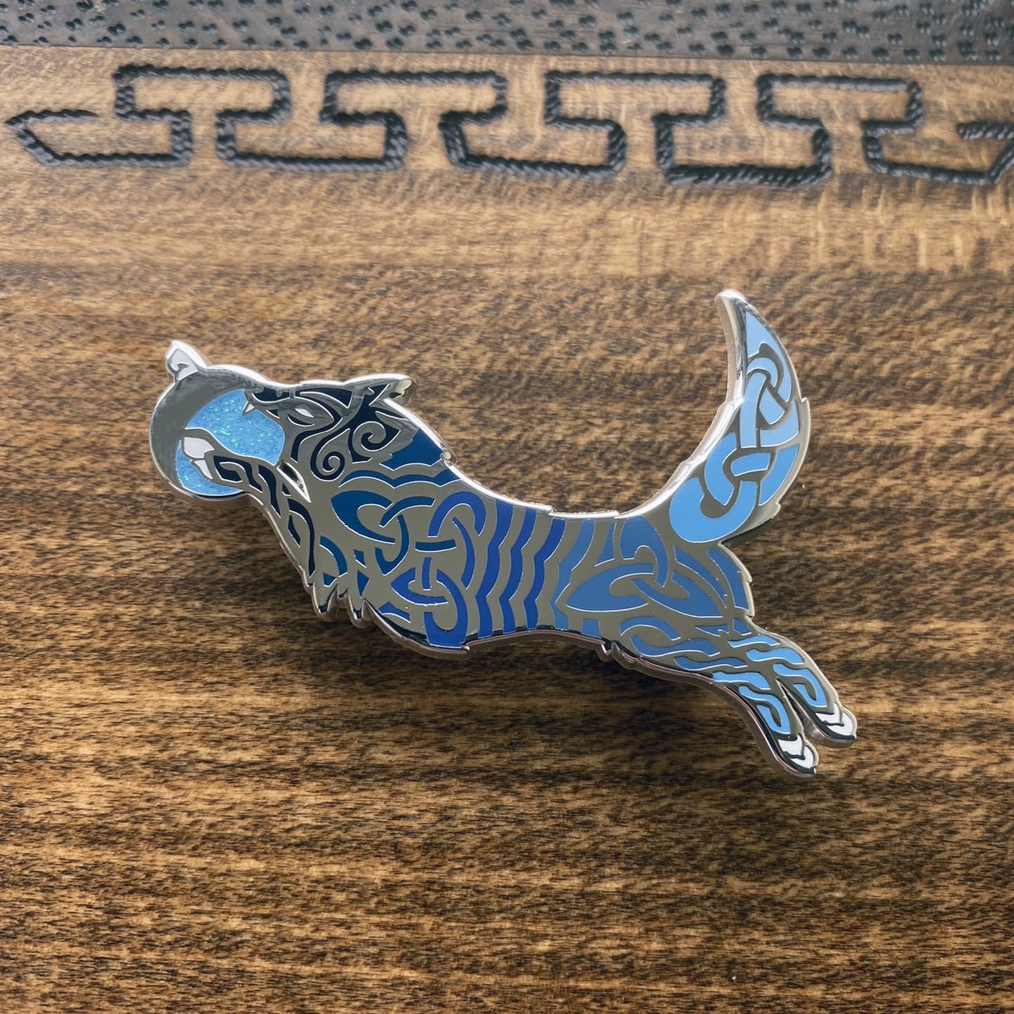 Enamel pin of the Norse icon Hati. Depicted as a wolf with a moon in its mouth. The pin contains knotwork with multiple blue enamel shades.