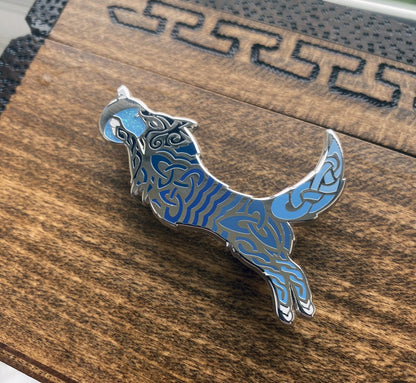 Enamel pin of the Norse icon Hati. Depicted as a wolf with a moon in its mouth. The pin contains knotwork with multiple blue enamel shades. Angle shows off silver plating.