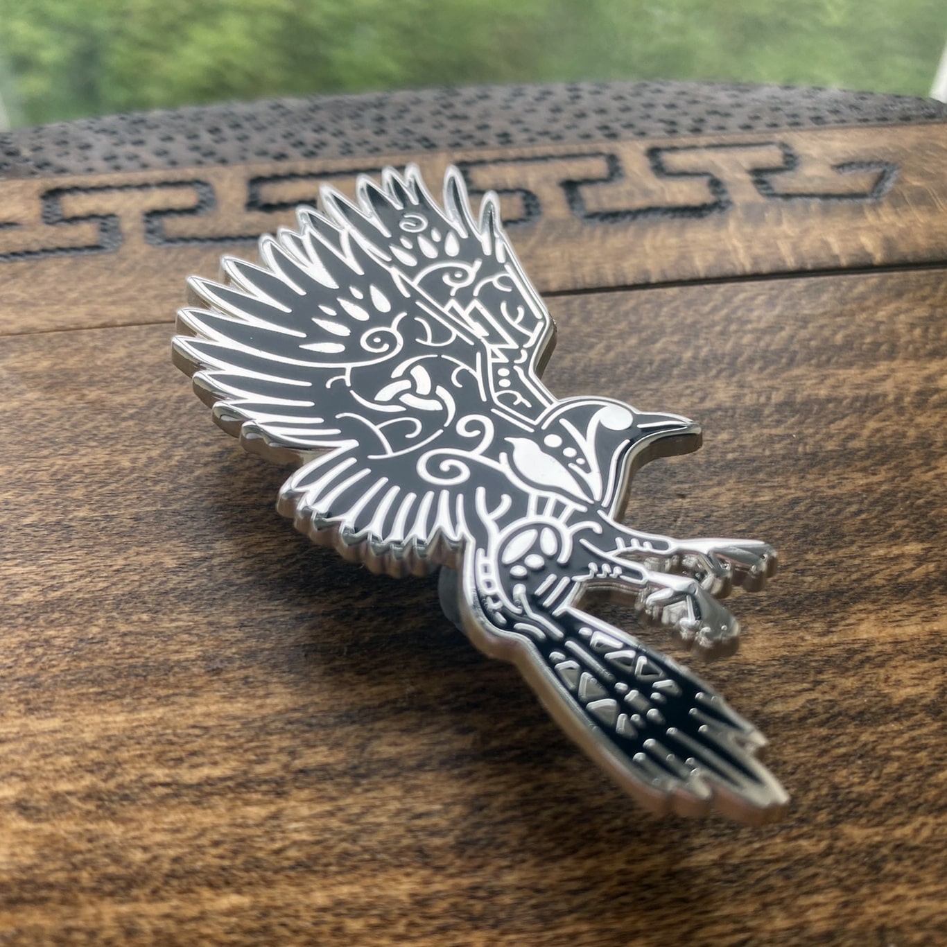 Norse god Odin, depicted as a raven enamel pin. The pin is silver plated and contains knotwork. Pin is at an angle to see the silver plating shine.