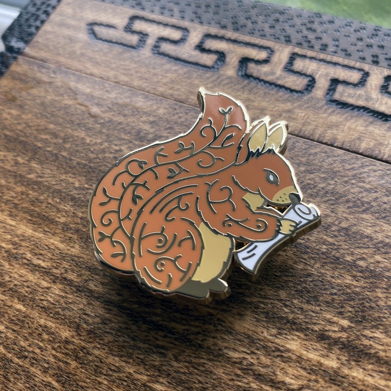 Gold-plated enamel pin of Norse icon Ratatoskr. The pin contains imagery of the world tree and is depicted as a squirrel holding a rolled up paper. Angle shows off gold-plating