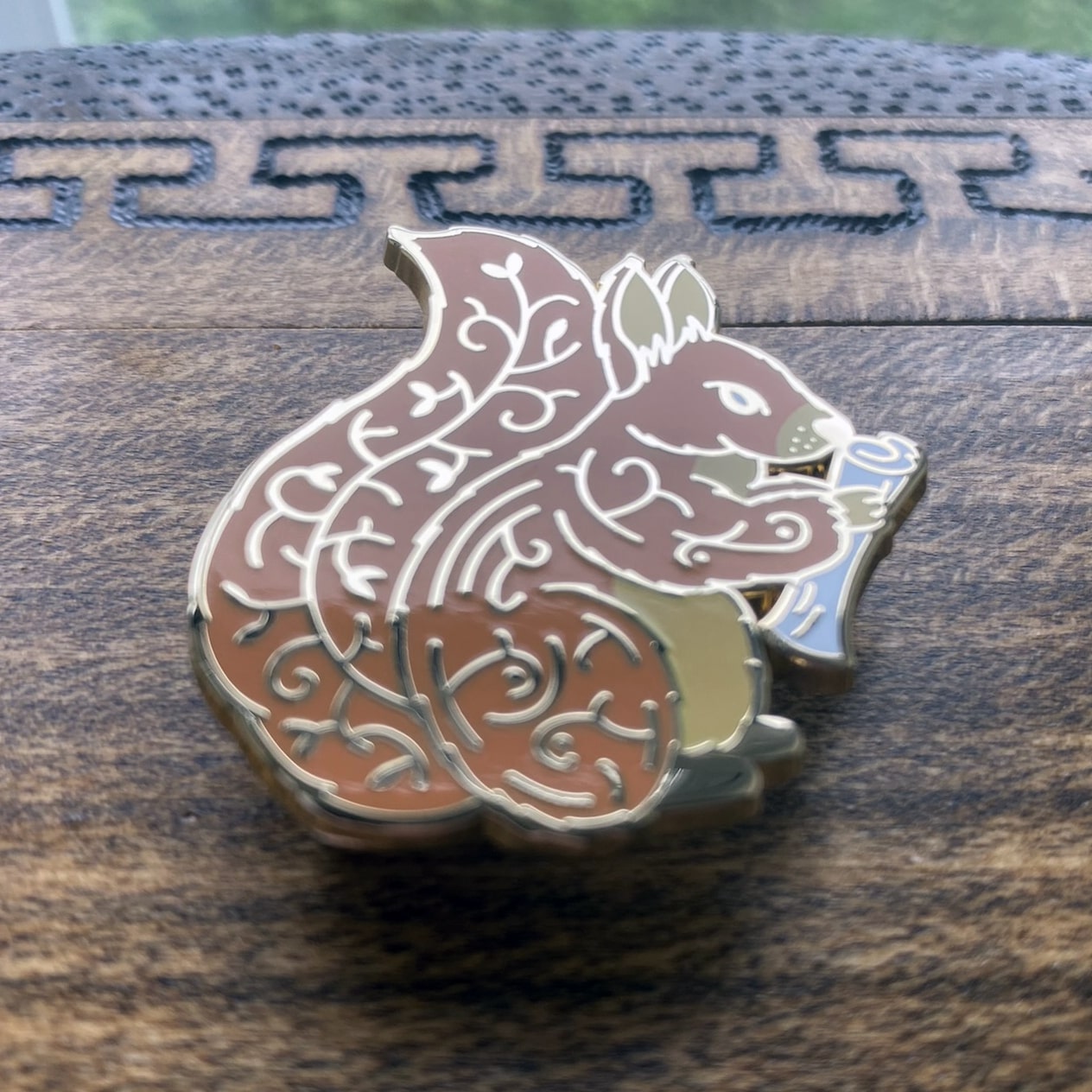 Gold-plated enamel pin of Norse icon Ratatoskr. The pin contains imagery of the world tree and is depicted as a squirrel holding a rolled up paper. Angle shows off gold-plating