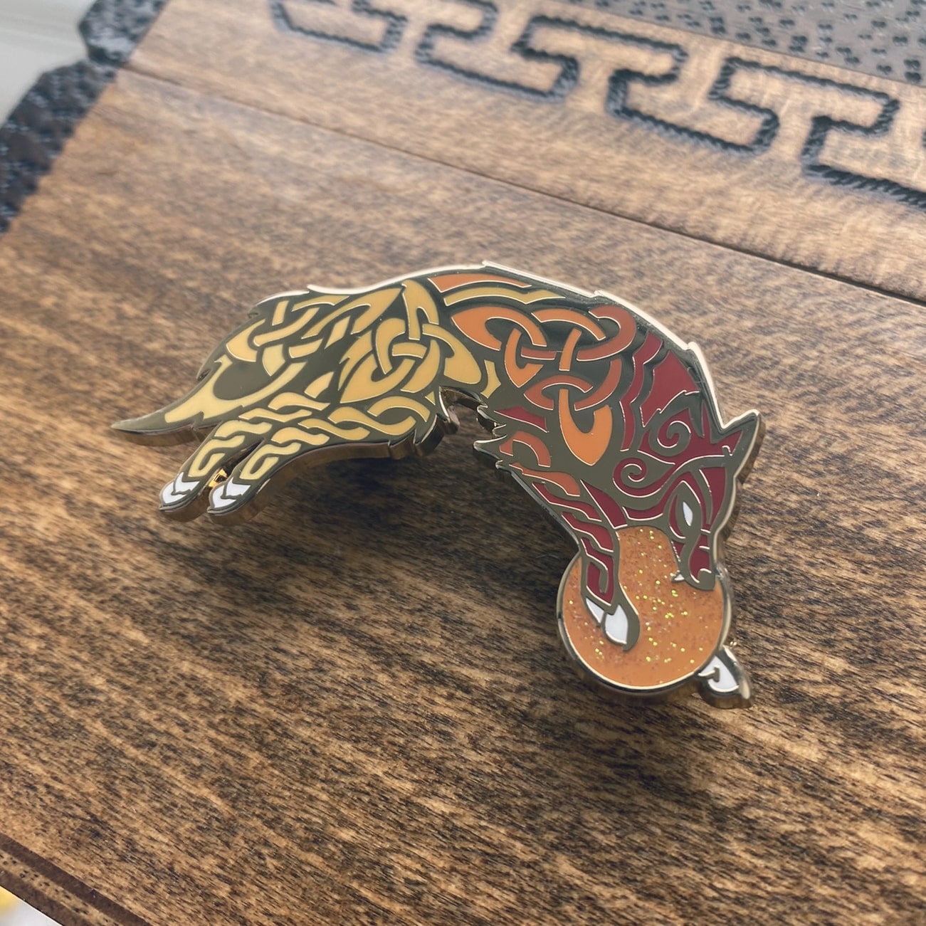 Gold-plated enamel pin of Skoll the Norse icon. The pin is a wolf and contains knotwork and multiple shades of red. Angle shows off gold-plating.